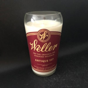 May include: A clear glass candle jar with a white candle inside. The jar has a red and gold label that reads "Weller The Original Wheated Bourbon Antique 107".