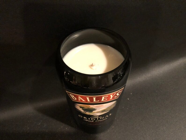 May include: A black glass candle holder with a white candle inside. The candle holder has a label that says "Baileys Original Irish Cream". The candle has a wooden wick.