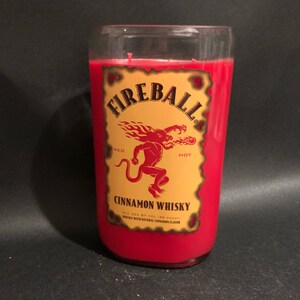 May include: A red candle in a glass jar with a Fireball Cinnamon Whisky label. The label features a red devil with flames and the words "Fireball", "Red Hot", and "Cinnamon Whisky".