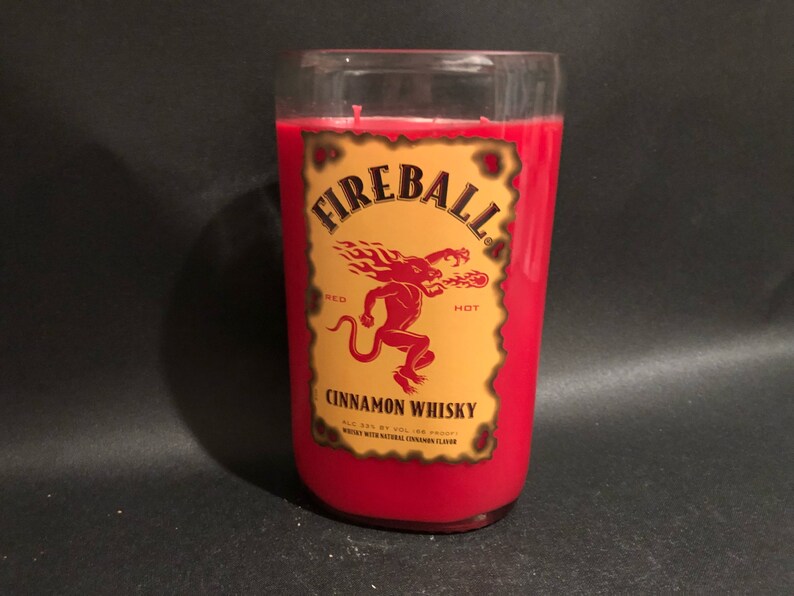 May include: A red candle in a glass jar with a Fireball Cinnamon Whisky label. The label features a red devil with flames and the words "Fireball", "Red Hot", and "Cinnamon Whisky".