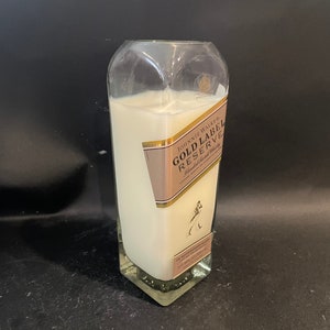 May include: A white candle in a clear glass bottle with a Johnnie Walker Gold Label Reserve label. The label is pink and gold with the text "Johnnie Walker Gold Label Reserve Blended Scotch Whisky".