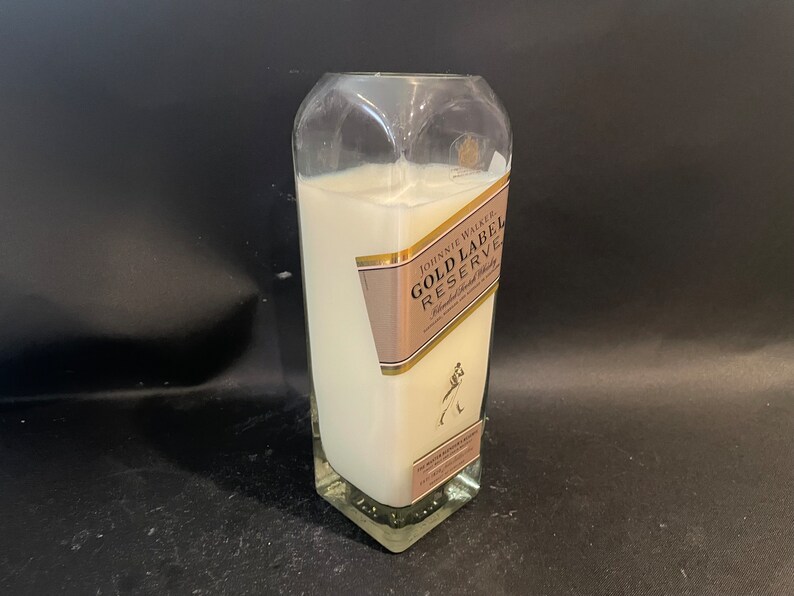 May include: A white candle in a clear glass bottle with a Johnnie Walker Gold Label Reserve label. The label is pink and gold with the text "Johnnie Walker Gold Label Reserve Blended Scotch Whisky".