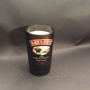 May include: A tall, dark glass with the Baileys Irish Cream logo, filled with a white liquid. The glass has a black and brown color scheme, with the Baileys logo prominently displayed. The glass is set against a dark background.