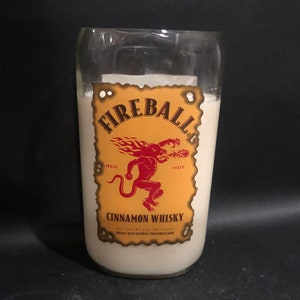 May include: A clear glass bottle candle with a white label featuring the Fireball Cinnamon Whisky logo. The logo is a red devil with flames and the words "Fireball", "Red Hot", and "Cinnamon Whisky".