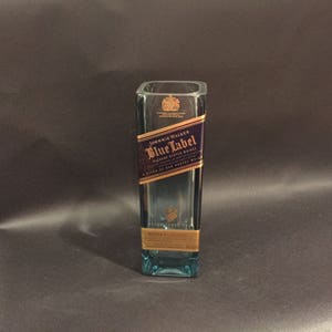 May include: A rectangular, clear glass bottle with a blue tint. The bottle features a label with the text "Johnnie Walker Blue Label" in gold and navy blue. The bottle is empty and stands upright.