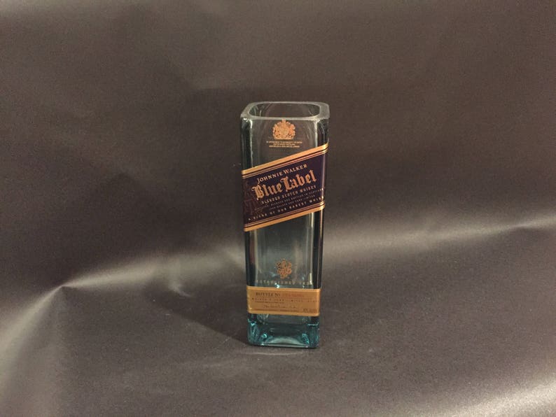 May include: A rectangular, clear glass bottle with a blue tint. The bottle features a label with the text "Johnnie Walker Blue Label" in gold and navy blue. The bottle is empty and stands upright.