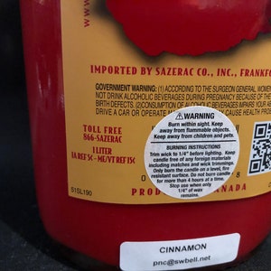 May include: A red glass bottle with a gold label. The label has the text "IMPORTED BY SAZERAC CO., INC., FRANKFORT, KY" and a warning about the consumption of alcoholic beverages. The bottle also has a QR code and the text "CINNAMON pnc@swbell.net".