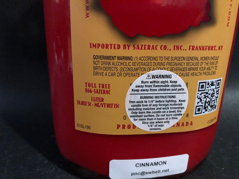 May include: A red glass bottle with a gold label. The label has the text "IMPORTED BY SAZERAC CO., INC., FRANKFORT, KY" and a warning about the consumption of alcoholic beverages. The bottle also has a QR code and the text "CINNAMON pnc@swbell.net".