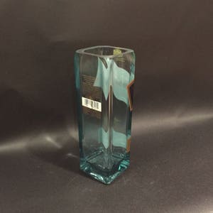 May include: A rectangular, clear glass vase with a light blue tint. The vase has a square opening and a label with a barcode. The vase is on a dark background.