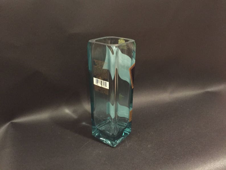 May include: A rectangular, clear glass vase with a light blue tint. The vase has a square opening and a label with a barcode. The vase is on a dark background.
