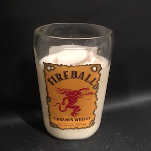 May include: A white candle in a clear glass jar with a Fireball Cinnamon Whisky label. The label is yellow with a red devil and the words "Fireball", "Red Hot", and "Cinnamon Whisky".