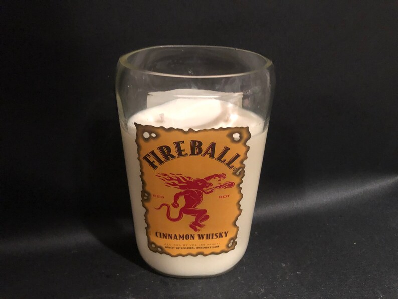 May include: A white candle in a clear glass jar with a Fireball Cinnamon Whisky label. The label is yellow with a red devil and the words "Fireball", "Red Hot", and "Cinnamon Whisky".
