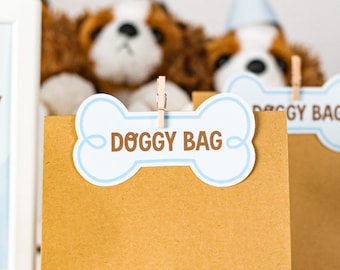 May include: Brown paper bags with blue and white bone-shaped tags that say "Doggy Bag". The bags are on a white table with blue and white dog-shaped cookies.