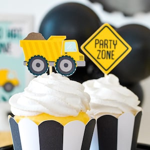 May include: Two cupcakes decorated with black and white striped cupcake liners. One cupcake has a yellow dumper truck topper and the other has a yellow sign that says "Party Zone".