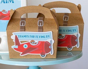 May include: A blue and white cake stand with two brown favor boxes decorated with red airplanes and the text "Thanks for flying by". The boxes are on a blue cake stand with a white base. There are also cookies in the shape of airplanes and clouds on a white plate.