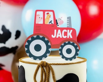 Könnte beinhalten: Ein weißer Kuchen, verziert mit schwarzen Kuhflecken und einer braunen Schleife aus Seil. Ein roter Traktor-Kuchentopper mit dem Namen "JACK" befindet sich oben auf dem Kuchen. Der Kuchen steht auf einem roten Kuchenständer.