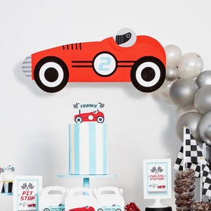 May include: A race car themed party table setting with a blue and white cake, red and black race car decorations, and silver balloons. The table is decorated with a banner that says "Two Fast".