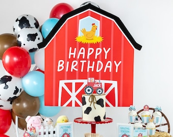 Puede incluir: Una mesa de fiesta de cumpleaños con temática de granja con un telón de fondo de granero rojo, un pastel decorado con manchas de vaca blancas y negras y una pancarta que dice "Feliz cumpleaños" y "Tres". La mesa está decorada con decoraciones con temática de animales de granja, incluyendo una vaca, una gallina y un tractor.