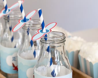 May include: Four clear glass bottles with blue and white striped straws and paper airplane propellers. The bottles have blue and white labels with red airplanes and the text "Jet Fuel". The bottles are sitting in a wooden tray.
