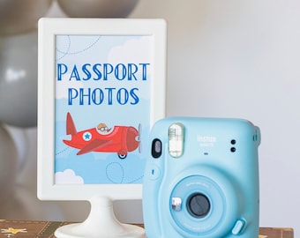 Może przedstawiać: Biały znak z niebieskimi i czerwonymi akcentami z napisem "Passport Photos" i ilustracją kreskówkowego samolotu. Niebieski aparat natychmiastowy stoi na brązowej walizce ze złotymi akcentami.