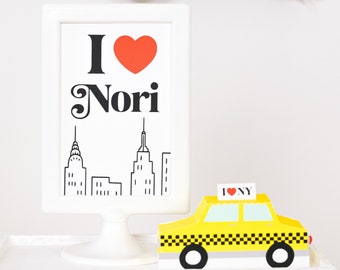 May include: A white framed sign with the text "I love Nori" in black and red. The sign features a black and white illustration of the New York City skyline. A yellow toy taxi cab with the text "I love NY" is in the foreground.