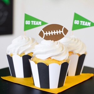 Può includere: Tre cupcakes con glassa bianca, involucri a righe bianche e nere e decorazioni a tema football americano. Ogni cupcake ha una bandiera verde con la scritta "GO TEAM" e un ritaglio di pallone da football marrone.