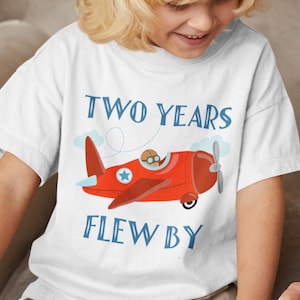 May include: A white t-shirt with a red aeroplane graphic and the text "TWO YEARS FLEW BY" in blue lettering.