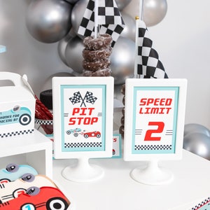 May include: A white table with a checkered racing theme party decor. There are two signs, one that says "Pit Stop" and the other that says "Speed Limit 2". There are also cookies shaped like race cars and a stack of chocolate donuts.