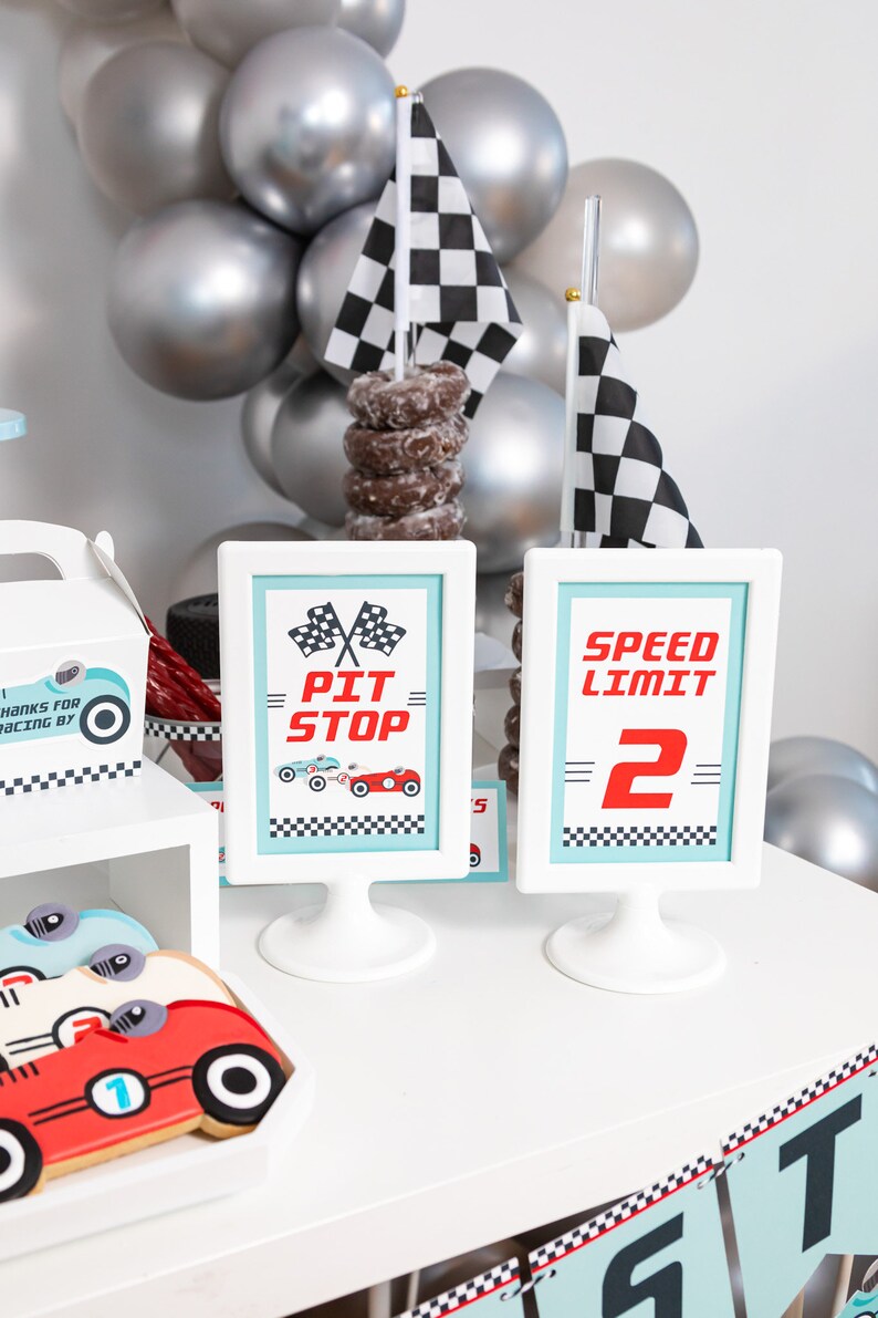 May include: A white table with a checkered racing theme party decor. There are two signs, one that says "Pit Stop" and the other that says "Speed Limit 2". There are also cookies shaped like race cars and a stack of chocolate donuts.