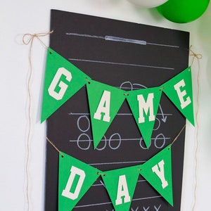 May include: Green and white paper banner with the words "GAME DAY" hanging on a black chalkboard with a football play drawn on it.