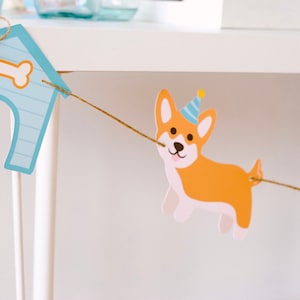 May include: A festive dog-themed banner featuring a corgi wearing a party hat, strung between two light blue dog houses with bone accents. The banner is made of paper and strung with twine, perfect for a birthday party.