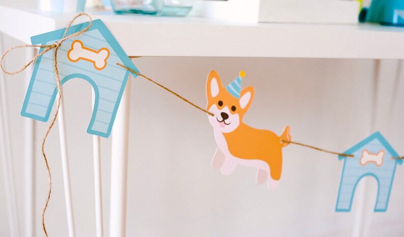 May include: A festive dog-themed banner featuring a corgi wearing a party hat, strung between two light blue dog houses with bone accents. The banner is made of paper and strung with twine, perfect for a birthday party.