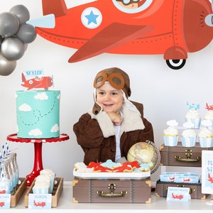 Op de afbeelding: Een verjaardagsfeestje met een luchtvaartthema. Een kind met een pilotenmuts en jas lacht voor een taart, cupcakes en vliegtuigdecoraties. Een banner leest "Happy Birthday!" en "Time Flies".