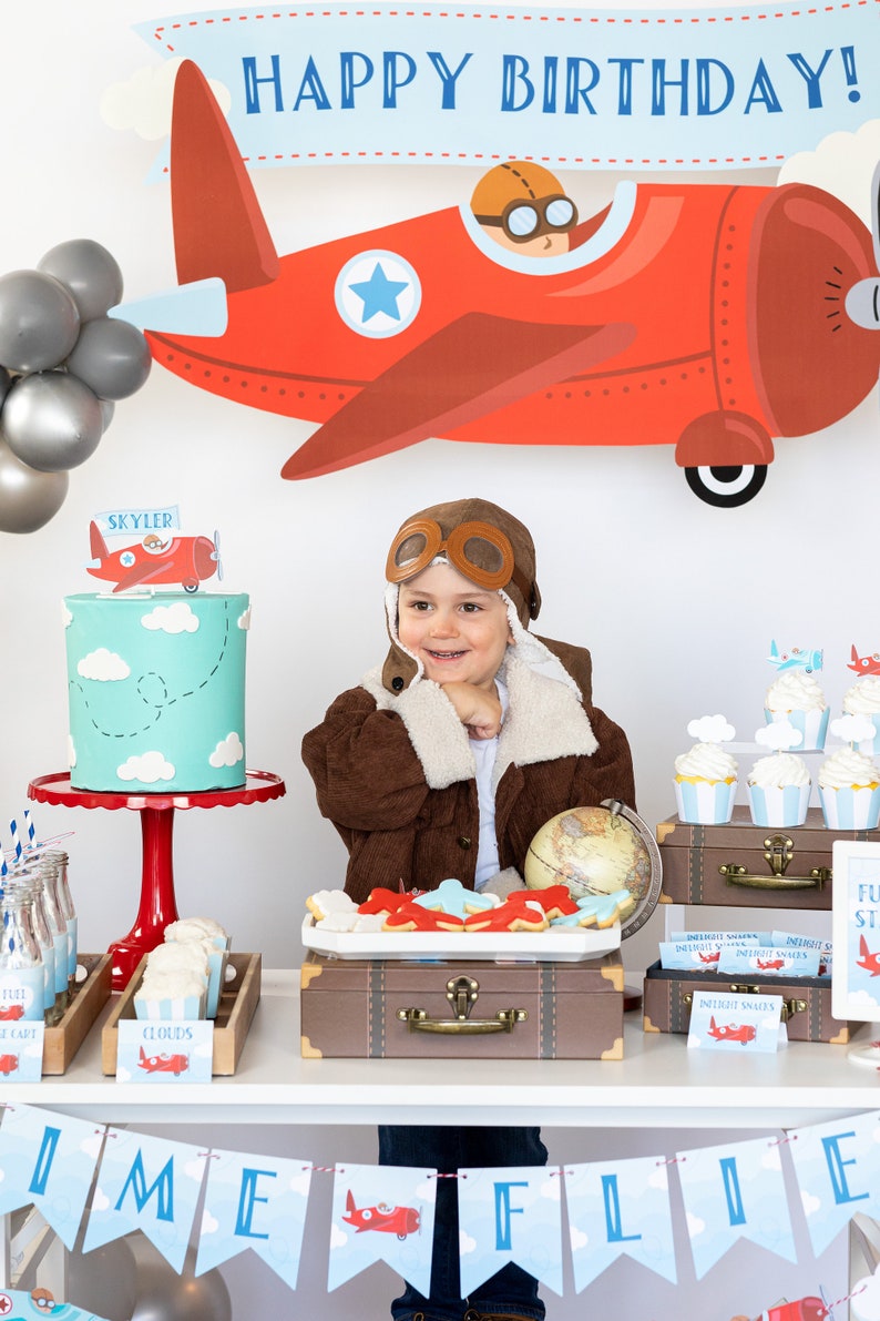 Op de afbeelding: Een verjaardagsfeestje met een luchtvaartthema. Een kind met een pilotenmuts en jas lacht voor een taart, cupcakes en vliegtuigdecoraties. Een banner leest "Happy Birthday!" en "Time Flies".