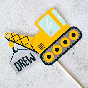 May include: A yellow construction vehicle with a black and gray track and a black hook. The vehicle is holding a gray block with the name "Drew" written on it.