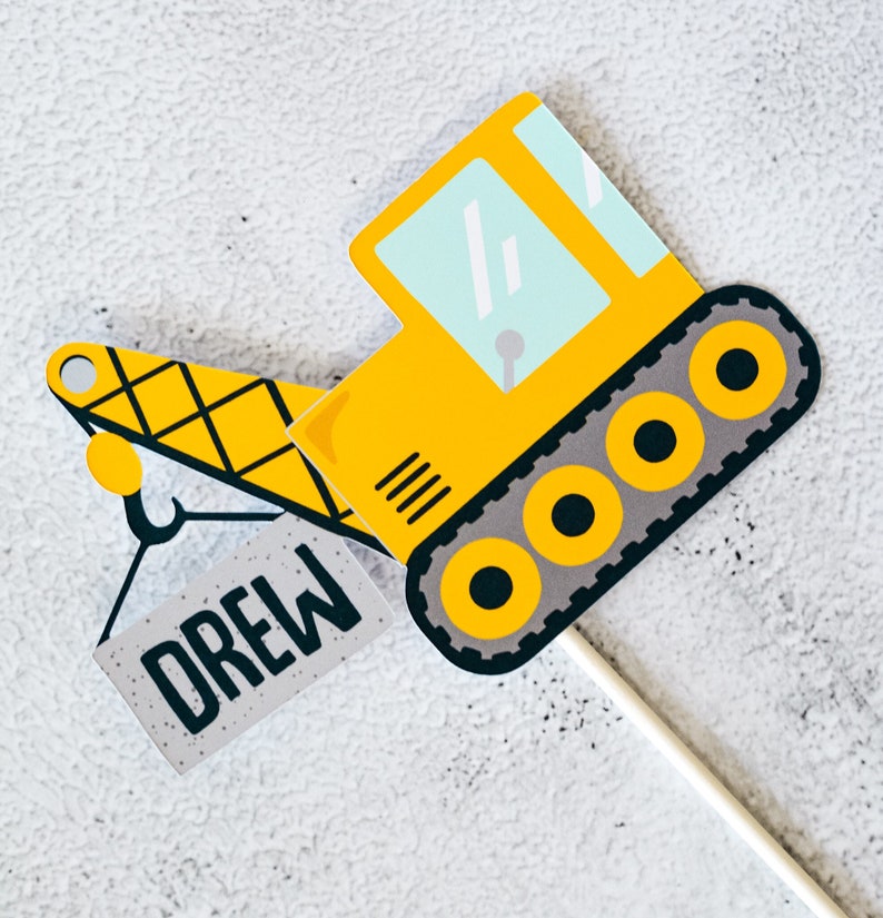 May include: A yellow construction vehicle with a black and gray track and a black hook. The vehicle is holding a gray block with the name "Drew" written on it.