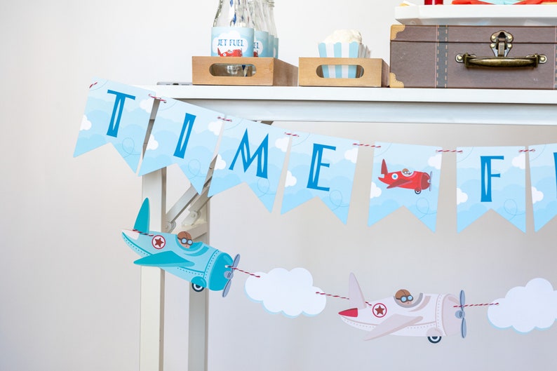 Op de afbeelding: Een feestelijke banner met de tekst "TIME FLIES" in blauwe letters tegen een hemelsblauwe achtergrond met wolken. Decoratieve vliegtuiguitsparingen in rood, lichtblauw en wit zijn aan de banner bevestigd.