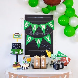 May include: A football-themed party table setting with green and white balloons, a chalkboard with the words "GAME DAY" written on it, a two-tiered cake decorated with footballs, and brown paper bags with footballs printed on them.