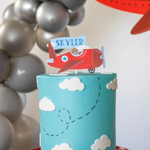 Puede incluir: Un pastel azul claro decorado con nubes blancas y un adorno para pastel de avi&oacute;n rojo con el nombre "Skyler". El pastel est&aacute; sobre un soporte rojo, con globos grises en el fondo. Un tema divertido para una fiesta de cumplea&ntilde;os.