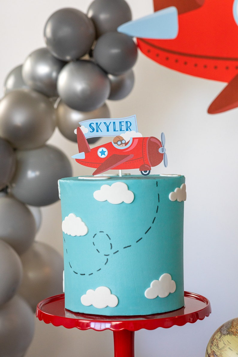 Puede incluir: Un pastel azul claro decorado con nubes blancas y un adorno para pastel de avi&oacute;n rojo con el nombre "Skyler". El pastel est&aacute; sobre un soporte rojo, con globos grises en el fondo. Un tema divertido para una fiesta de cumplea&ntilde;os.
