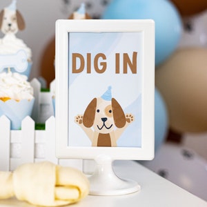 May include: A white framed sign with the words "DIG IN" in brown letters. Below the text is a cartoon dog wearing a party hat. The sign is on a white stand, with a bone and cupcakes in the background.