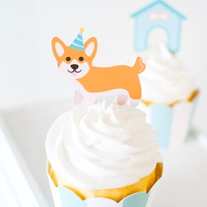 May include: A cupcake with white frosting and a dog-shaped topper wearing a blue and yellow party hat. The cupcake is in a blue and white striped paper liner.