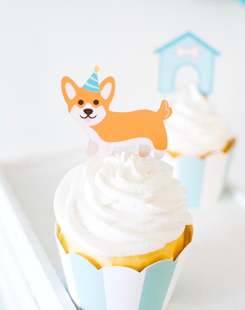 May include: A cupcake with white frosting and a dog-shaped topper wearing a blue and yellow party hat. The cupcake is in a blue and white striped paper liner.