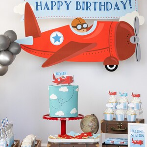 Puede incluir: Decoraci&oacute;n de fiesta de cumplea&ntilde;os con tem&aacute;tica de avi&oacute;n. Una gran decoraci&oacute;n de avi&oacute;n rojo con una pancarta que dice "HAPPY BIRTHAY!" se muestra encima de un pastel, cupcakes y otras golosinas. El pastel es azul claro con nubes blancas.