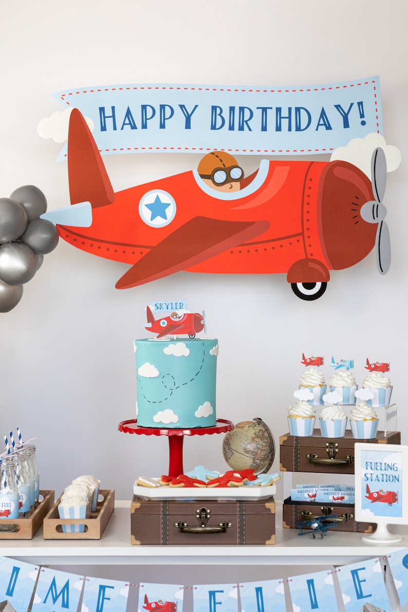 Puede incluir: Decoraci&oacute;n de fiesta de cumplea&ntilde;os con tem&aacute;tica de avi&oacute;n. Una gran decoraci&oacute;n de avi&oacute;n rojo con una pancarta que dice "HAPPY BIRTHAY!" se muestra encima de un pastel, cupcakes y otras golosinas. El pastel es azul claro con nubes blancas.