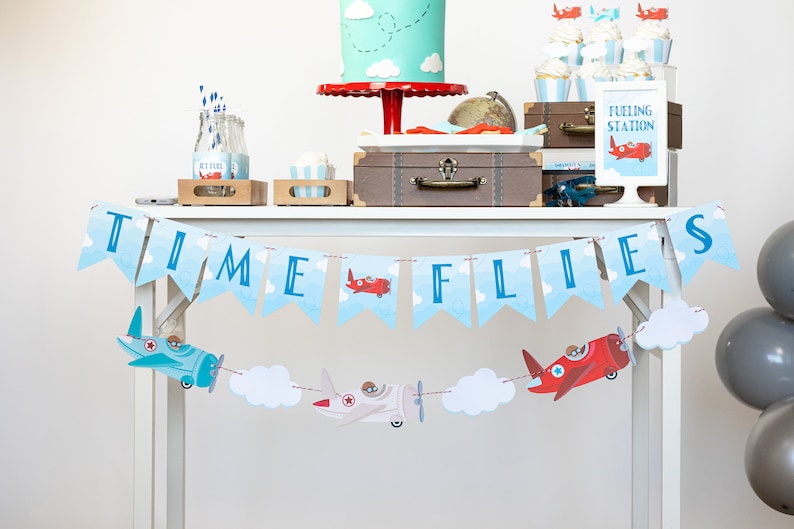 Op de afbeelding: Een feesttafel versierd met een luchtvaartthema. Een banner leest "TIME FLIES" met vliegtuig- en wolkuitsparingen. De tafel bevat een taart, cupcakes en een bord met de tekst "Fueling Station".