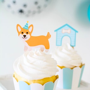 May include: Two cupcakes with white frosting and blue and white striped cupcake liners. One cupcake has a paper dog wearing a party hat and the other has a paper doghouse.