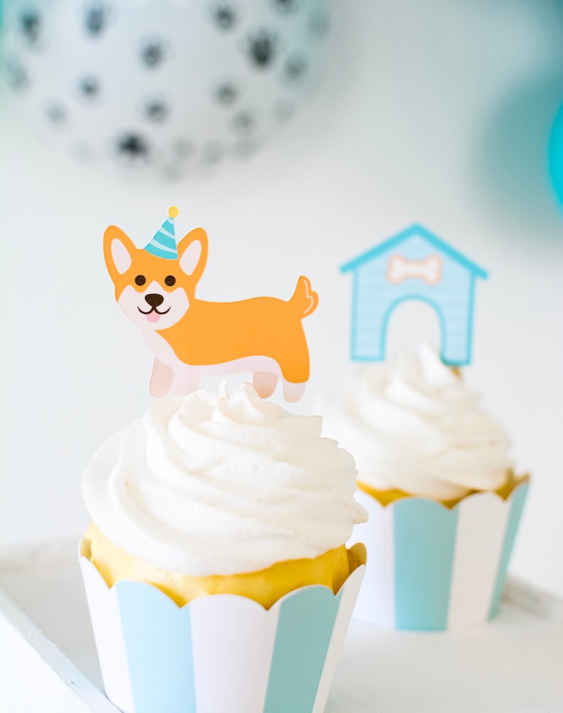 May include: Two cupcakes with white frosting and blue and white striped cupcake liners. One cupcake has a paper dog wearing a party hat and the other has a paper doghouse.