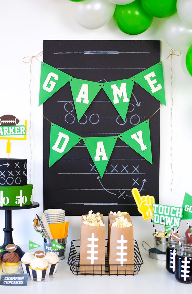 May include: Green and white 'Game Day' banner with a football field drawn on a black chalkboard. There are brown paper bags filled with popcorn, cupcakes with football designs, and a football-themed cake.