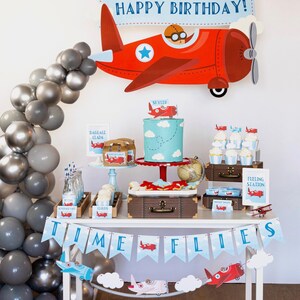 Puede incluir: Decoraci&oacute;n de fiesta de cumplea&ntilde;os con tem&aacute;tica de aviaci&oacute;n. Un gran avi&oacute;n rojo con un piloto est&aacute; encima de una mesa con pastel, cupcakes y otras golosinas. Una pancarta dice "Happy Birthday!" y un arco de globos.